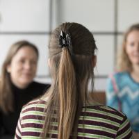 En pige sidder overfor to kvinder i en samtale, hvor man kun kan se hende bagfra. 